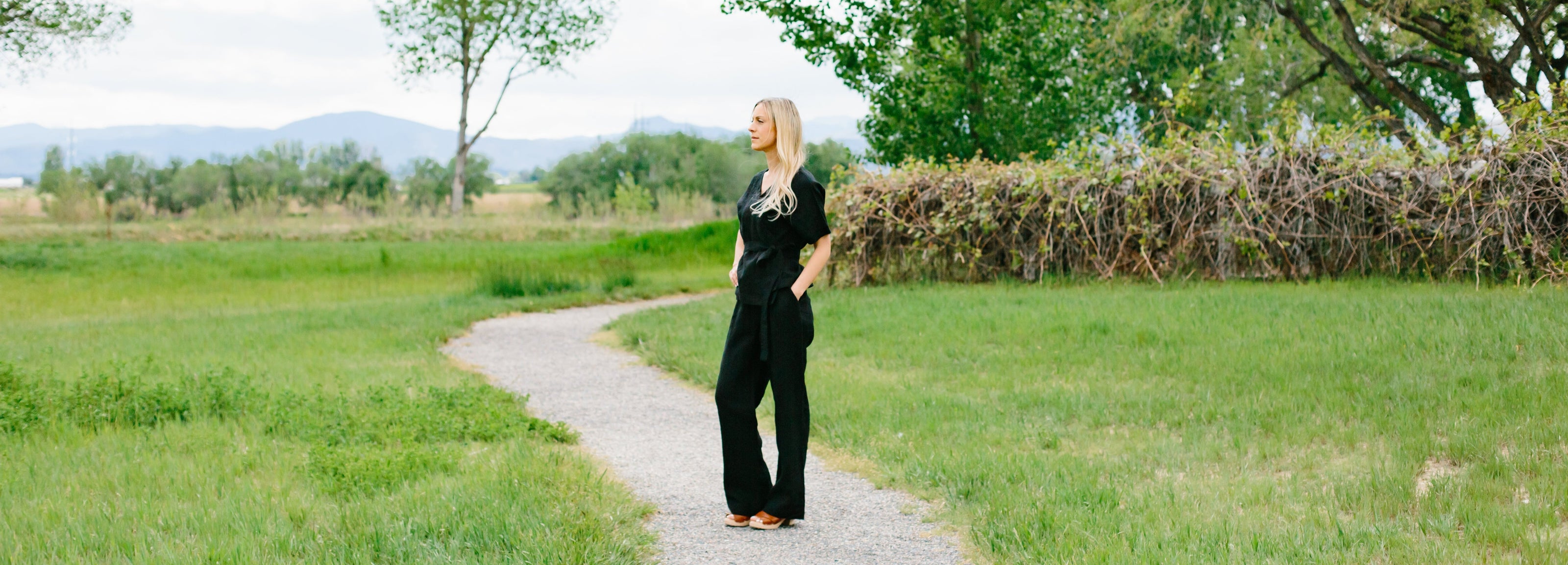 Woman standing on a path in black linen wrap top and black linen tailored pants in a green field with trees and mountains in the background